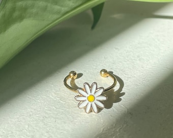 May include: A gold-colored ring with a white daisy flower design and a yellow center. The ring has a split shank design with small gold balls at the ends. The ring is set against a white surface with a green leaf in the background.
