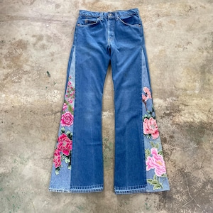 May include: Blue denim jeans with a split design featuring embroidered pink and red flowers. The jeans have a classic five-pocket design and a button and zip closure. The floral embroidery adds a unique, artistic touch to the garment.