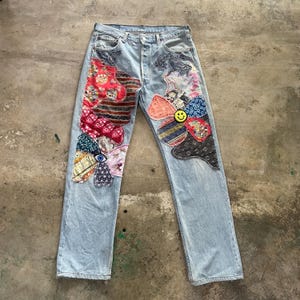 Può includere: Jeans di denim blu chiaro con motivi floreali patchwork colorati. Le toppe includono vari modelli e trame, con una faccina sorridente gialla al centro di un fiore. I jeans hanno un classico design a cinque tasche.