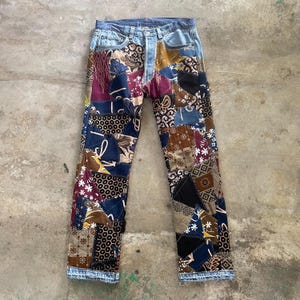 Può includere: Jeans a figura intera caratterizzati da un design patchwork composto da vari quadrati di tessuto nelle tonalità del blu, marrone, nero e bordeaux. Le toppe presentano motivi diversi, tra cui disegni floreali, geometrici e a nido d'ape.