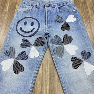 May include: Light blue denim jeans with a large smiley face patch and multiple four-leaf clover designs in shades of black, white, and blue. The jeans have a classic five-pocket design and a button and zipper closure.