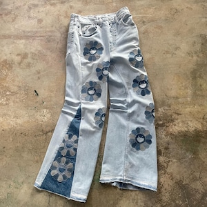 May include: Light wash denim jeans with a flared leg design. The jeans are adorned with multiple blue and gray flower appliqués. The jeans are laid flat on a concrete surface.