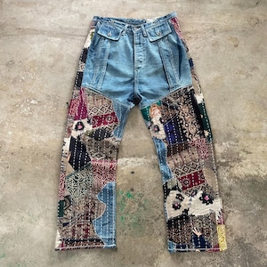 May include: High-waisted denim pants featuring a light blue wash upper section with two front flap pockets and a button fly. The lower legs are constructed from a patchwork of various multicolored fabric squares with visible white stitching.