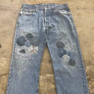 Könnte beinhalten: Hellblaue Jeans mit dekorativen Aufnähern und weißen Nähten. Die Aufnäher haben verschiedene Formen, darunter Kreise und ein Friedenszeichen. Die Jeans hat einen Knopfverschluss und ein klassisches Fünf-Taschen-Design.