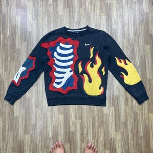 May include: A navy blue sweatshirt with a distinctive design featuring red, white, and yellow flame and bone-like shapes. The Nike logo is visible on the chest. The sweatshirt has long sleeves and a crew neck.