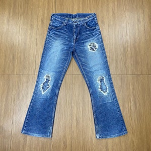 May include: Blue denim jeans with distressed patches on the knees. The jeans have a classic five-pocket design and a flared leg opening. The denim has a faded wash, giving it a worn look.