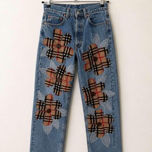 Op de afbeelding: Lichtblauwe spijkerbroek met grote bloemenapplicaties. De bloemen zijn gemaakt van een bruine, beige en rode geruite stof. Witte bladvormige details zijn rond de bloemen gestikt. De jeans hangt aan een metalen hanger.