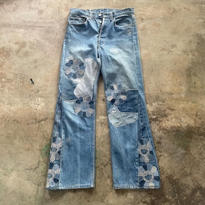 Puede incluir: Vaqueros de mezclilla azul claro con parches de flores en tonos azules y grises. Los vaqueros tienen bragueta con botones y un diseño clásico de cinco bolsillos. Los parches de flores están en la parte delantera y los lados de las piernas.