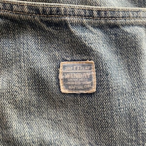 May include: Close-up of denim fabric with a rectangular patch. The patch has a light blue border and features text including "SAFETY FIRST" and "WINDAY". The denim is a faded blue with visible stitching.