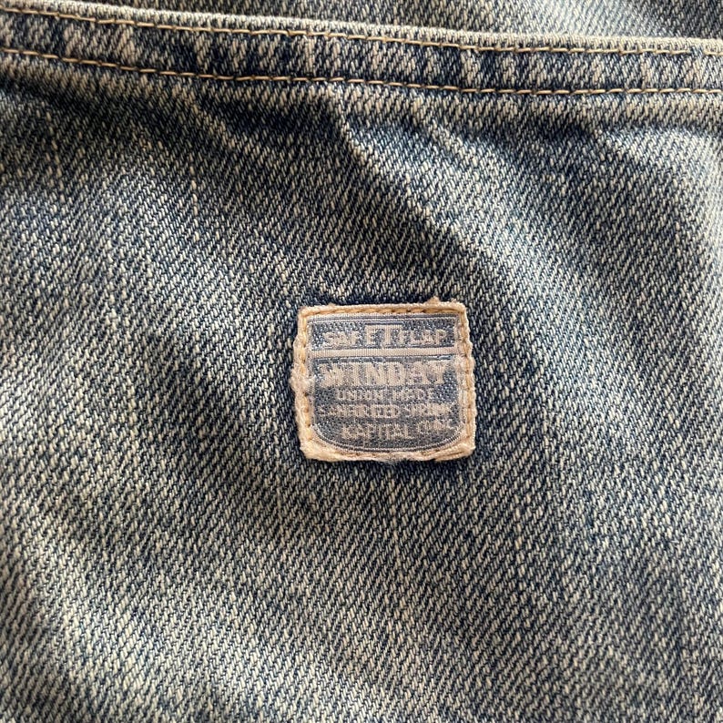 May include: Close-up of denim fabric with a rectangular patch. The patch has a light blue border and features text including "SAFETY FIRST" and "WINDAY". The denim is a faded blue with visible stitching.