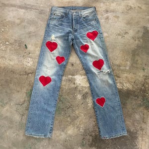 May include: Light blue denim jeans with distressed details and red heart patches. The jeans have a classic five-pocket design and a button-fly closure. The red hearts are sewn onto the front of the jeans.