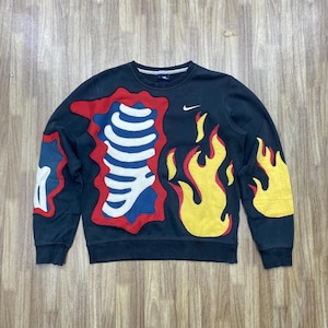 May include: Black crewneck sweatshirt with a unique design. Features a skeleton-like graphic in white, red, and blue, along with yellow flame accents. The Nike logo is visible on the chest. A stylish and eye-catching piece.