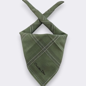 May include: An olive green handkerchief featuring a plaid design and the text "Christian Dior" in black. The handkerchief is folded into a triangle shape, with the ends crossed over each other.