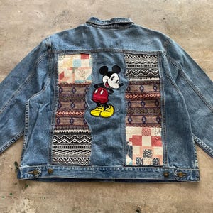 Könnte beinhalten: Hellblaue Jeansjacke mit Patchwork-Details und einem Mickey Mouse-Patch. Die Rückseite der Jacke zeigt farbenfrohe Stoffbahnen mit geometrischen und floralen Mustern sowie eine große Mickey Mouse Applikation.