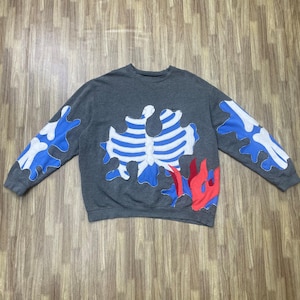 May include: A grey sweatshirt with a distinctive design. It features white and blue bone-like shapes on the sleeves and chest, and red and blue flame-like shapes at the bottom. The sweatshirt is made of a soft material.