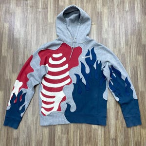 May include: Grey hooded sweatshirt with a distinctive design. It features red and white shapes, resembling a rib cage, on one side and blue flames on the other. The sweatshirt has a drawstring hood and long sleeves.