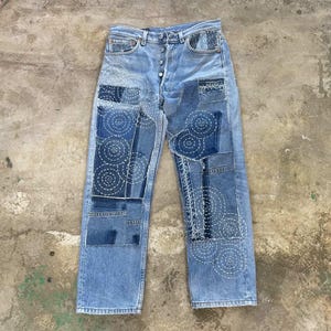 May include: Light blue denim jeans with multiple patches of varying shades of blue. The patches are adorned with white, circular stitching. The jeans have a button fly and a classic five-pocket design.