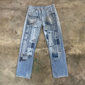 May include: Light blue denim jeans with patchwork and visible stitching details. The jeans feature various denim patches and decorative stitching, creating a unique, distressed look. The jeans have a classic five-pocket design and a button closure.