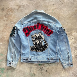 May include: A light blue denim jacket with a large embroidered patch on the back that says "Just  Bint" in black and red lettering. The patch features a black and white illustration of two people in a boat. The jacket has two green patches on the shoulders.