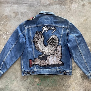 Könnte beinhalten: Blaue Jeansjacke mit großem gesticktem Adlerdesign auf dem Rücken, darüber das Wort "Japan". Die Jacke hat ein klassisches Knopfdesign und ist aus mittelblauem Denim gefertigt.