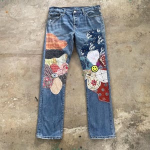 Può includere: Jeans in denim blu con grandi disegni patchwork irregolari sulla parte anteriore di entrambe le gambe. Le toppe sono costituite da vari motivi in tessuto, tra cui stampe floreali, geometriche e a onde, con una piccola icona gialla a faccina sorridente.