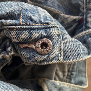May include: Close-up of a light blue denim jacket with a silver button. The button is embossed with the text "KAPITAL BRAND". The jacket has visible stitching and a worn appearance.
