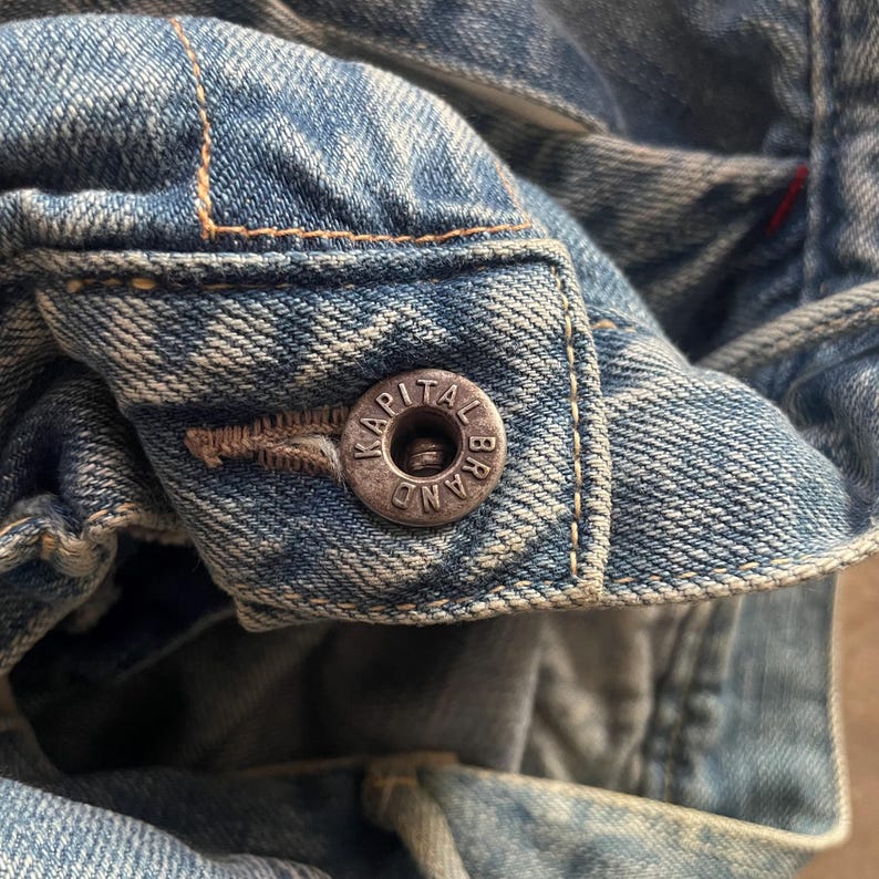 May include: Close-up of a light blue denim jacket with a silver button. The button is embossed with the text "KAPITAL BRAND". The jacket has visible stitching and a worn appearance.
