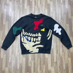 Op de afbeelding: Zwart sweatshirt met ronde hals en een skeletontwerp. Het sweatshirt heeft een grote, gebroken witte skeletafbeelding op de voorkant, met kleurrijke patches in rood, groen, geel en wit. Het Adidas-logo is zichtbaar op de borst.