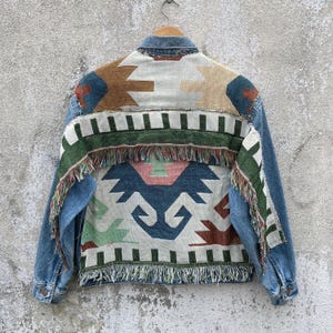 May include: A blue denim jacket featuring a back panel made of woven fabric with a geometric pattern in shades of cream, blue, green, brown, and pink. The woven section includes horizontal bands with decorative fringe along the edges and hem.