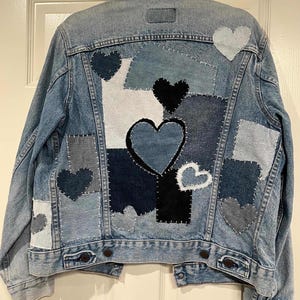 May include: A light blue denim jacket with a patchwork design. The back of the jacket features heart-shaped patches in various shades of blue and white. The patches are of different sizes and textures, creating a unique and artistic look.