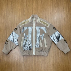 May include: A beige fleece zip-up jacket featuring the word FARNESE in bold letters on the shoulders. The front displays large silver metallic quilted panels and lettering. Each sleeve includes a silver quilted section and a circular patch.