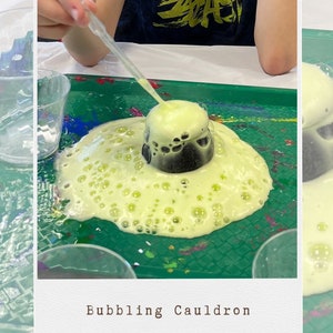 May include: A green tray with a black cauldron bubbling over with yellow foam. The foam is overflowing the cauldron and spreading across the tray. A person is using a dropper to add more liquid to the cauldron.
