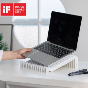 May include: A white laptop stand with a laptop on top of it. The stand is made of a series of angled, white slats. The laptop is closed and has a black screen. The stand is on a white table with two black pens and a white coffee cup on a saucer.