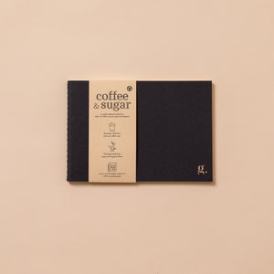 May include: A black notebook with a brown paper cover that says "coffee & sugar" and has a small illustration of a coffee cup. The notebook has a gold "J.G." logo on the front cover.
