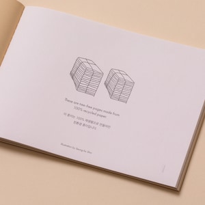 May include: A close-up of a page from a book with a black and white illustration of two stacks of paper. The text reads "These are tree-free pages made from 100% recycled paper. 이 종이는 100% 재생프로들어진 친환경 종이랍니다. Illustration by Seung Ho Shie"