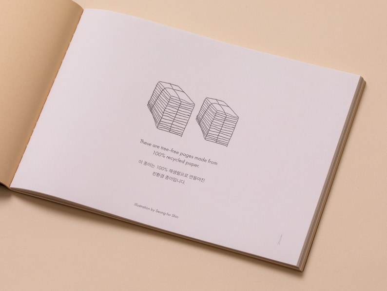 May include: A close-up of a page from a book with a black and white illustration of two stacks of paper. The text reads "These are tree-free pages made from 100% recycled paper. 이 종이는 100% 재생프로들어진 친환경 종이랍니다. Illustration by Seung Ho Shie"