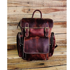 May include: A brown leather backpack with two large front pockets and a top handle. The backpack has two adjustable straps and a zipper closure.