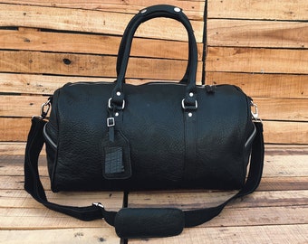 Può includere: Una borsa da viaggio in pelle nera con una tracolla e un'etichetta per i bagagli. La borsa ha accessori in argento e una chiusura a zip.