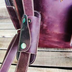 May include: Close-up of a brown leather backpack strap with white stitching and metal accents. The strap has a flap with two metal snaps.