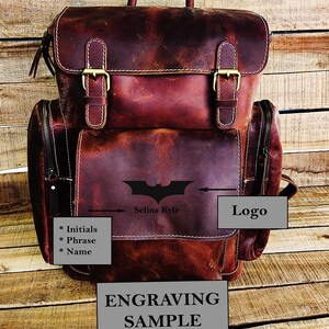 May include: A brown leather backpack with a zippered front pocket and two adjustable straps. The backpack has a Batman logo engraved on the front with the text "Selina Kyle" below it. The text "Logo" is written in a white box on the right side of the image. The text "Engraving Sample" is written in a white box at the bottom of the image.