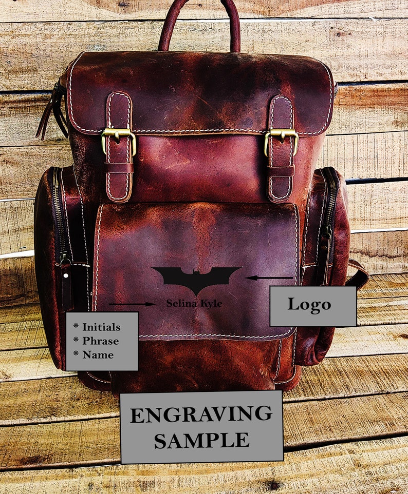 May include: A brown leather backpack with a zippered front pocket and two adjustable straps. The backpack has a Batman logo engraved on the front with the text "Selina Kyle" below it. The text "Logo" is written in a white box on the right side of the image. The text "Engraving Sample" is written in a white box at the bottom of the image.