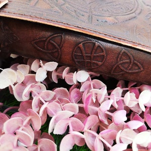 K&ouml;nnte beinhalten: Nahaufnahme eines braunen Lederjournals mit keltischen Knoten und einem gepr&auml;gten Kreisdesign auf dem Einband. Das Journal liegt auf einem Bett aus rosa und wei&szlig;en Eukalyptusbl&auml;ttern.