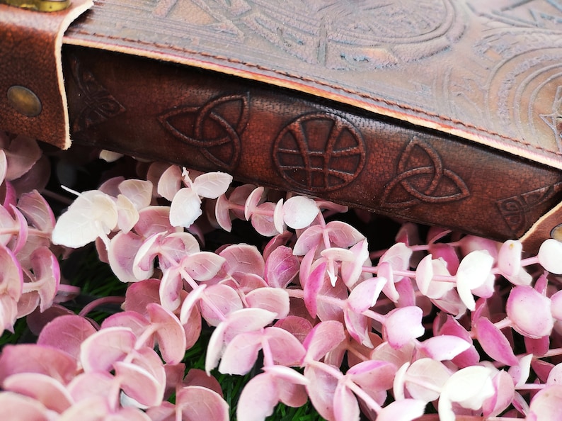 K&ouml;nnte beinhalten: Nahaufnahme eines braunen Lederjournals mit keltischen Knoten und einem gepr&auml;gten Kreisdesign auf dem Einband. Das Journal liegt auf einem Bett aus rosa und wei&szlig;en Eukalyptusbl&auml;ttern.