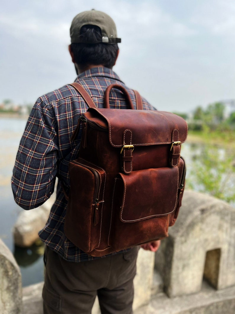 May include: A brown leather backpack with a large front pocket and two adjustable straps. The backpack has a zipper closure and brass buckles.