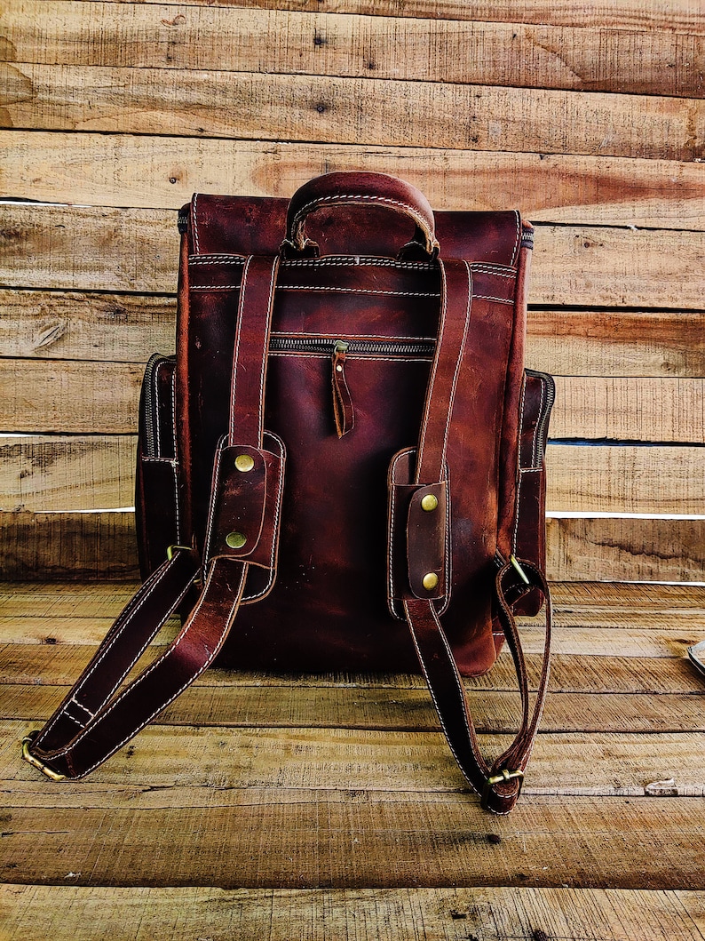 May include: A brown leather backpack with adjustable straps and a zippered front pocket. The backpack has a top handle and brass hardware.