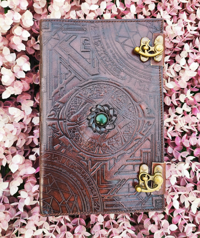 K&ouml;nnte beinhalten: Ein braunes Lederjournal mit aufwendigen gepr&auml;gten Designs und einem Akzent aus gr&uuml;nem Edelstein. Das Journal hat zwei Messingverschl&uuml;sse.