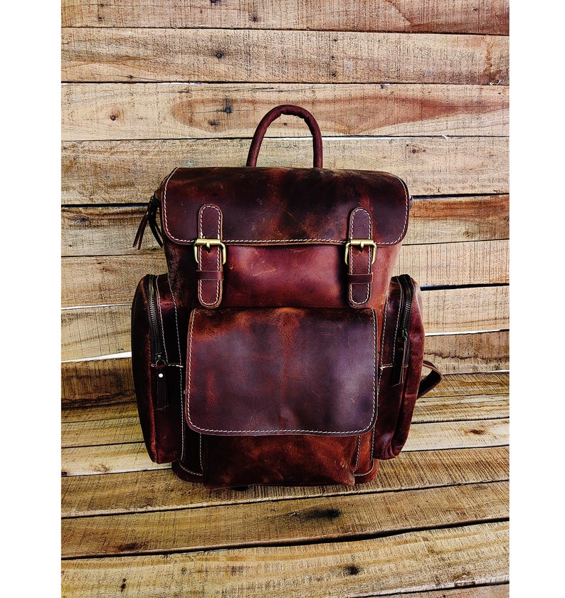 May include: A brown leather backpack with two large front pockets and a top handle. The backpack has two adjustable straps and a zipper closure.