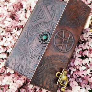 K&ouml;nnte beinhalten: Ein braunes Lederjournal mit aufwendigen gepr&auml;gten Designs und einem gr&uuml;nen Edelsteinschloss. Das Journal hat einen Klappverschluss mit einem Metallriegel.