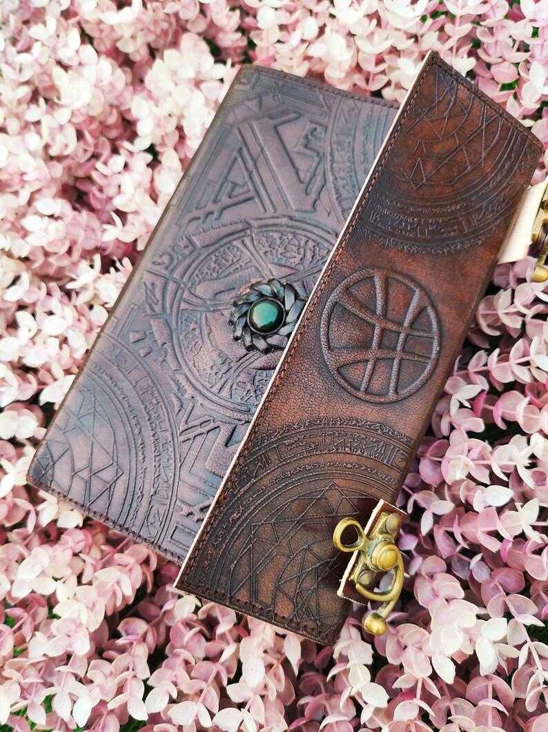 K&ouml;nnte beinhalten: Ein braunes Lederjournal mit aufwendigen gepr&auml;gten Designs und einem gr&uuml;nen Edelsteinschloss. Das Journal hat einen Klappverschluss mit einem Metallriegel.