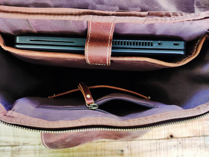 May include: A brown leather backpack with a zippered pocket and a flap that opens to reveal a padded laptop compartment. The laptop compartment is lined with a soft, dark fabric.